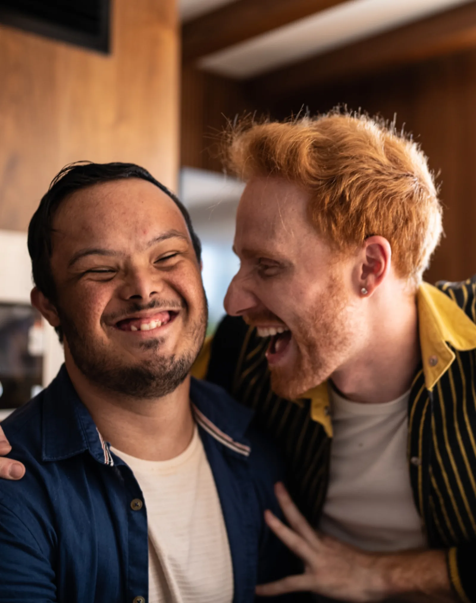 Two young men laughing warmly together at home, representing the inclusive friendship and disability support Noon Care provides to NDIS participants
